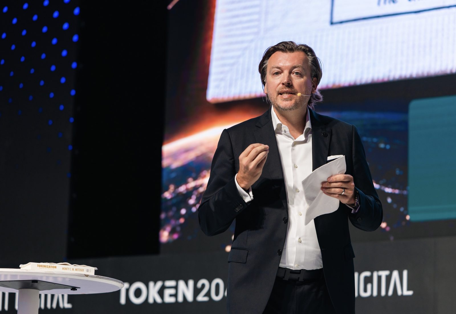 Monty Metzger, Founder and Chairman of Toto Finance, speaking at TOKEN2049 Singapore 2026 with a copy of Tokenization: Capital for Humanity on the lectern