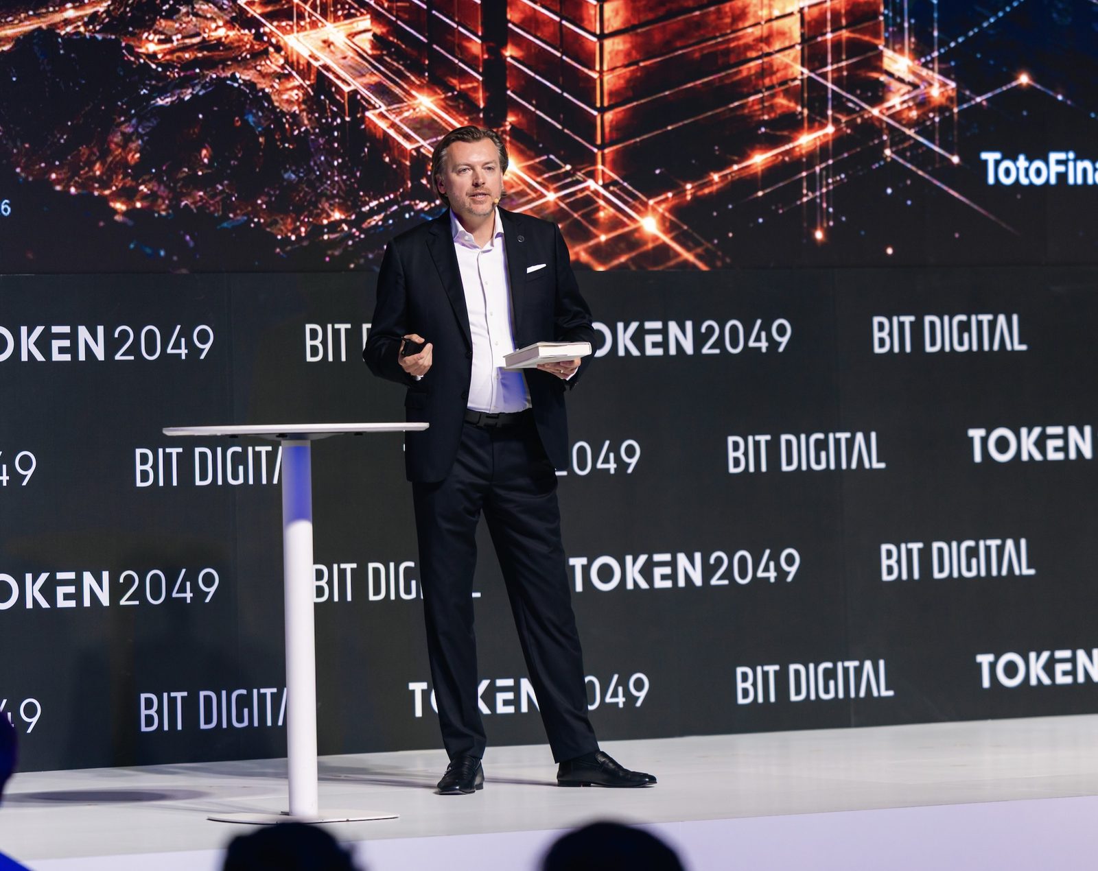 Monty Metzger speaking at TOKEN2049 Singapore 2026 holding his book Tokenization: Capital for Humanity, with the Toto Finance keynote slide behind him